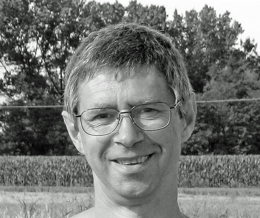 Smiling middle-aged man with glasses, head-and-shoulders portrait outdoors with trees and a field behind him.