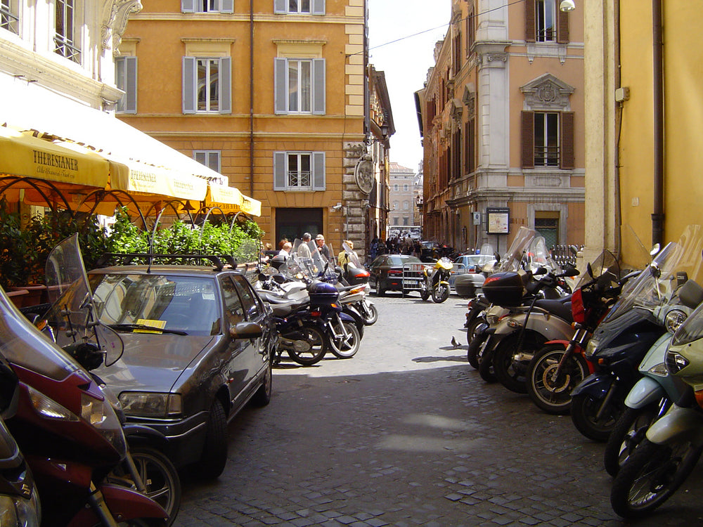 Cobblestone street in a European city, lined with parked scooters and cars, yellow café awnings and warm ochre buildings.