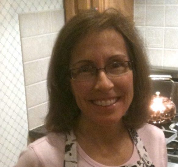 Smiling woman wearing glasses and a patterned apron standing in a tiled kitchen