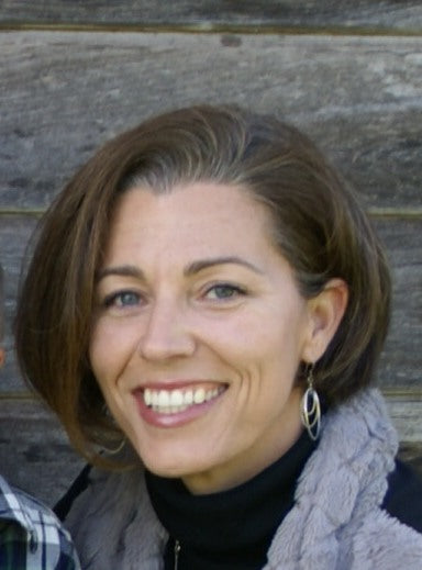 Smiling woman with short brown hair wearing a gray scarf and black top, posed in front of a weathered wooden wall