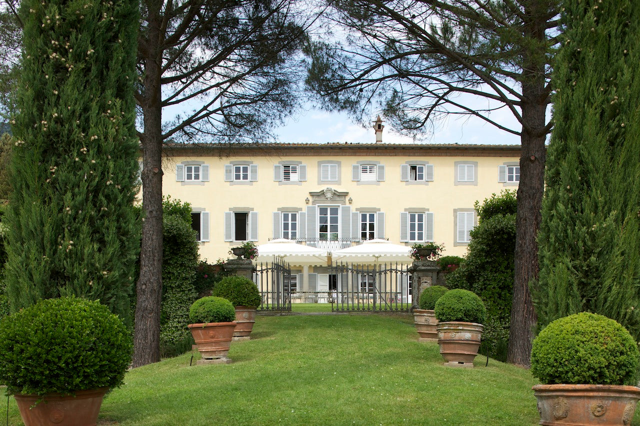 Pale-yellow Italian villa with shuttered windows and gated terrace, framed by cypress trees and potted boxwood.