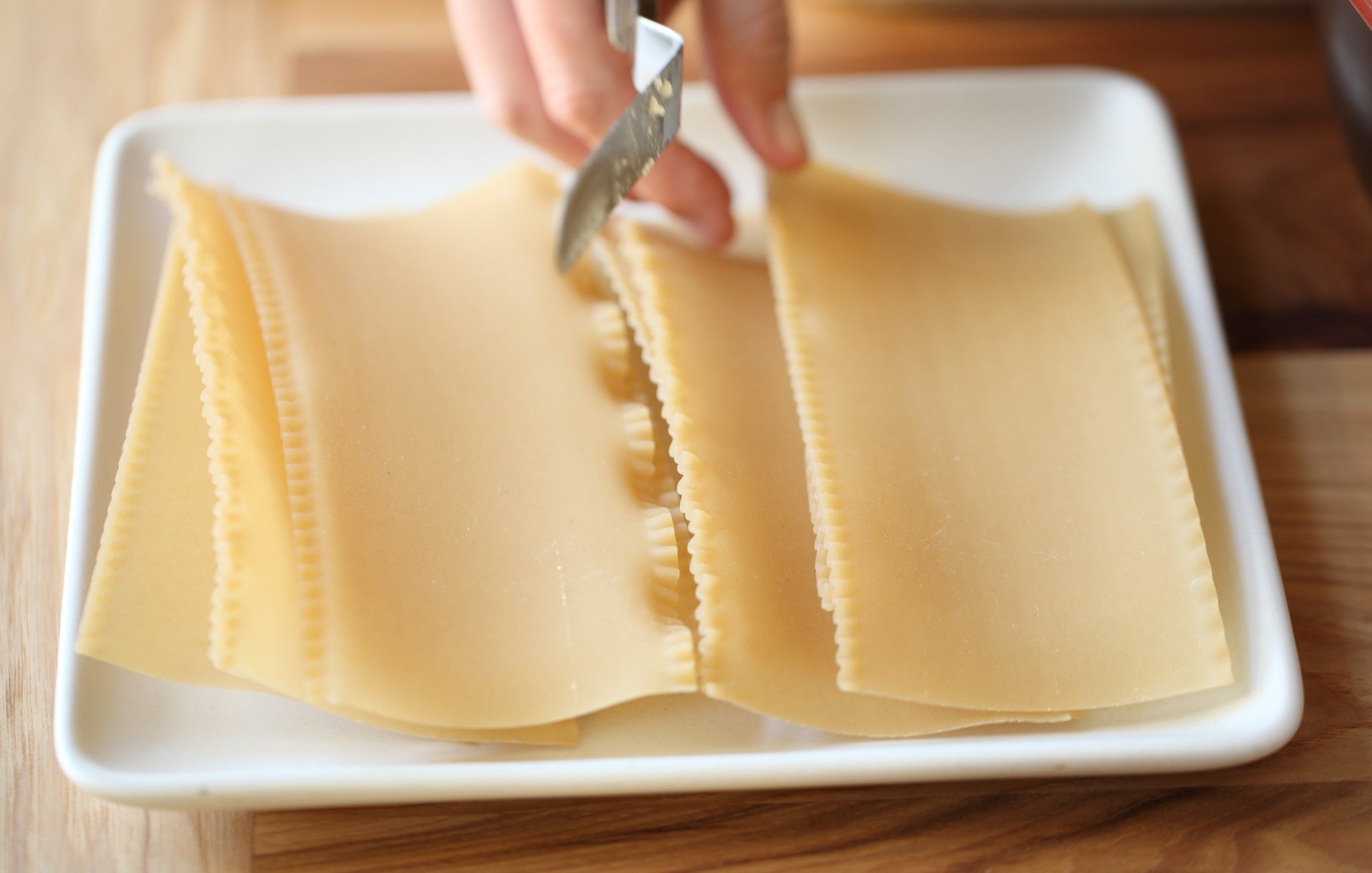 Uncooked lasagna pasta sheets on a white plate, a hand trimming the ruffled edges with a knife