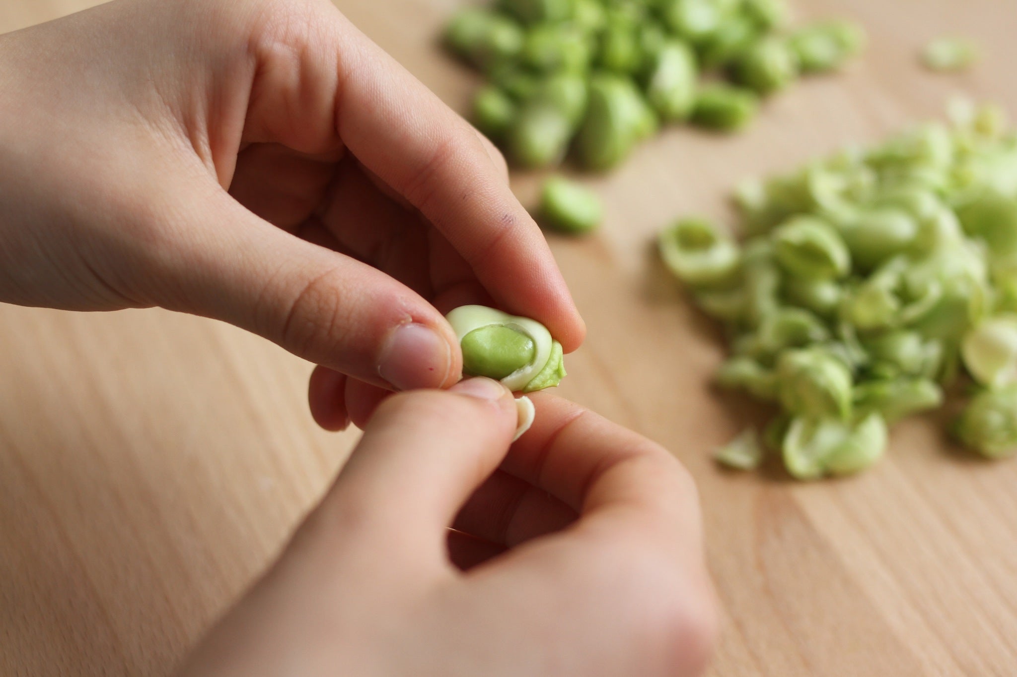 Hands removing a bright green fava bean from its pod with more shelled beans on a wooden surface.