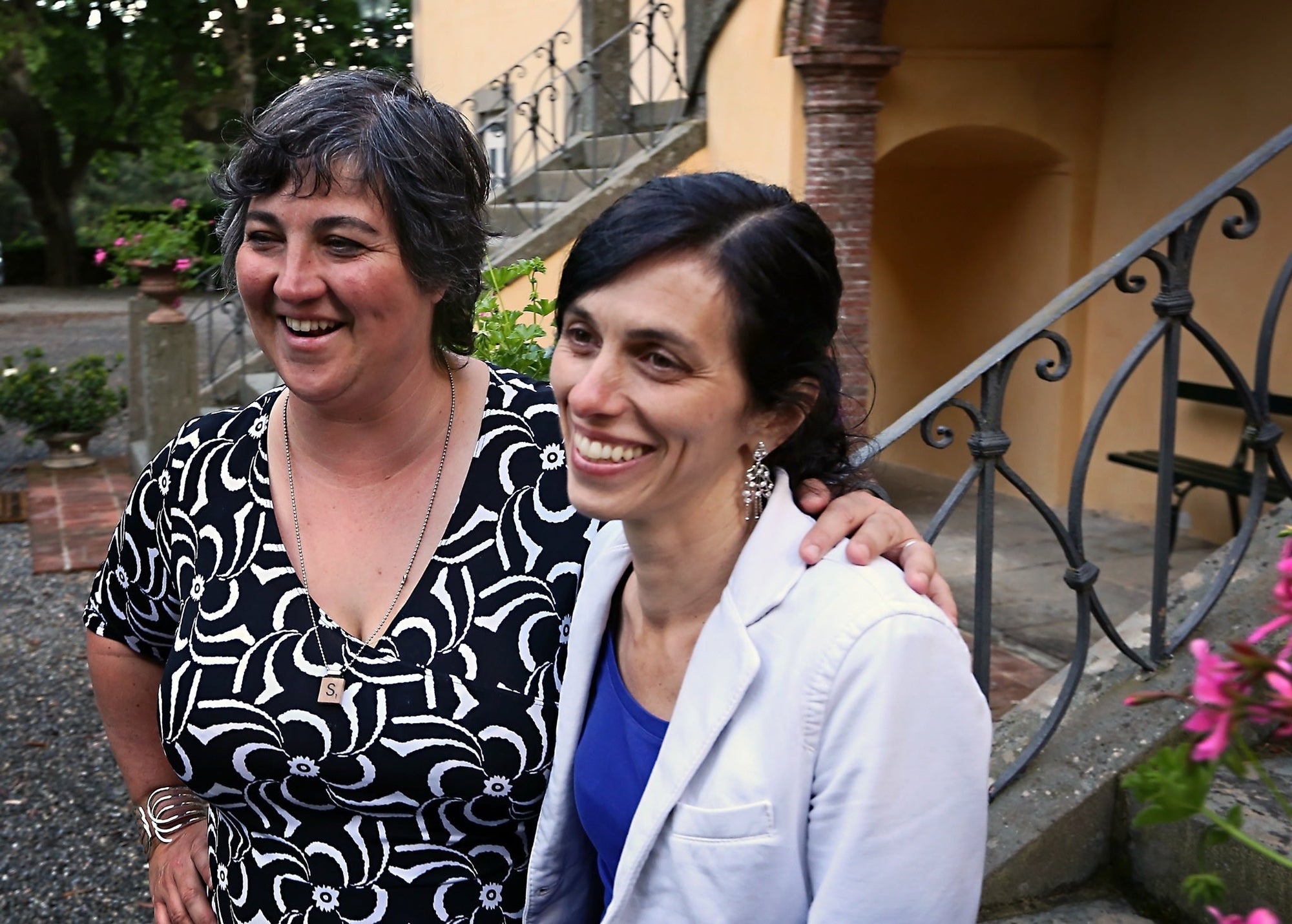 Two women smiling together by stone steps and wrought-iron railing; necklace pendant reads 'S'