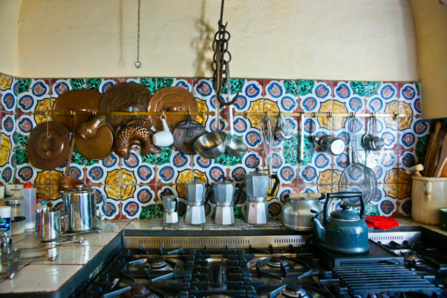 Italian style stove and gas burners with colorful patterned tile backsplash, hanging copper pans, three moka pots, and a black kettle.