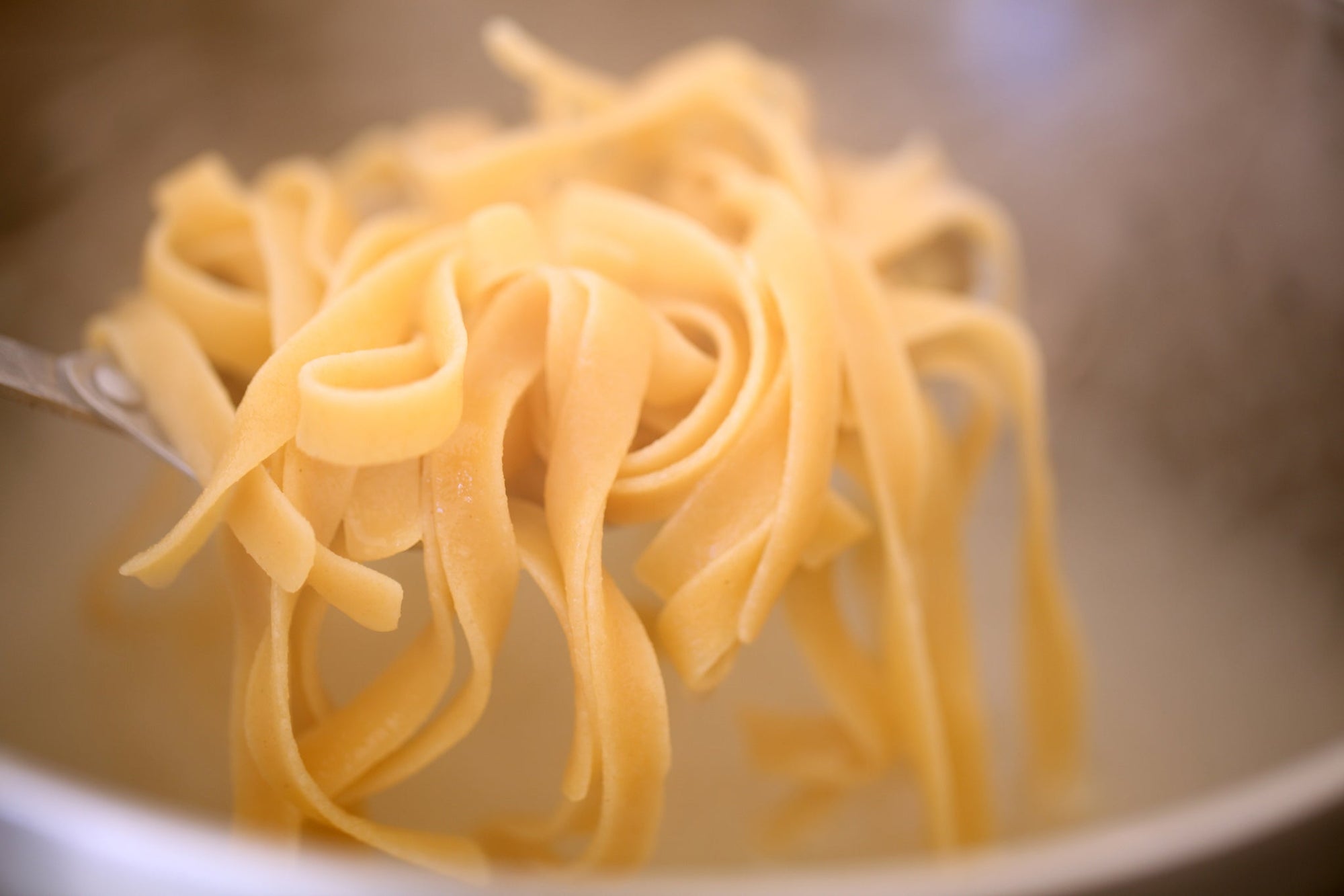 Fork lifting fresh tagliatelle-style pasta strands above cooking water