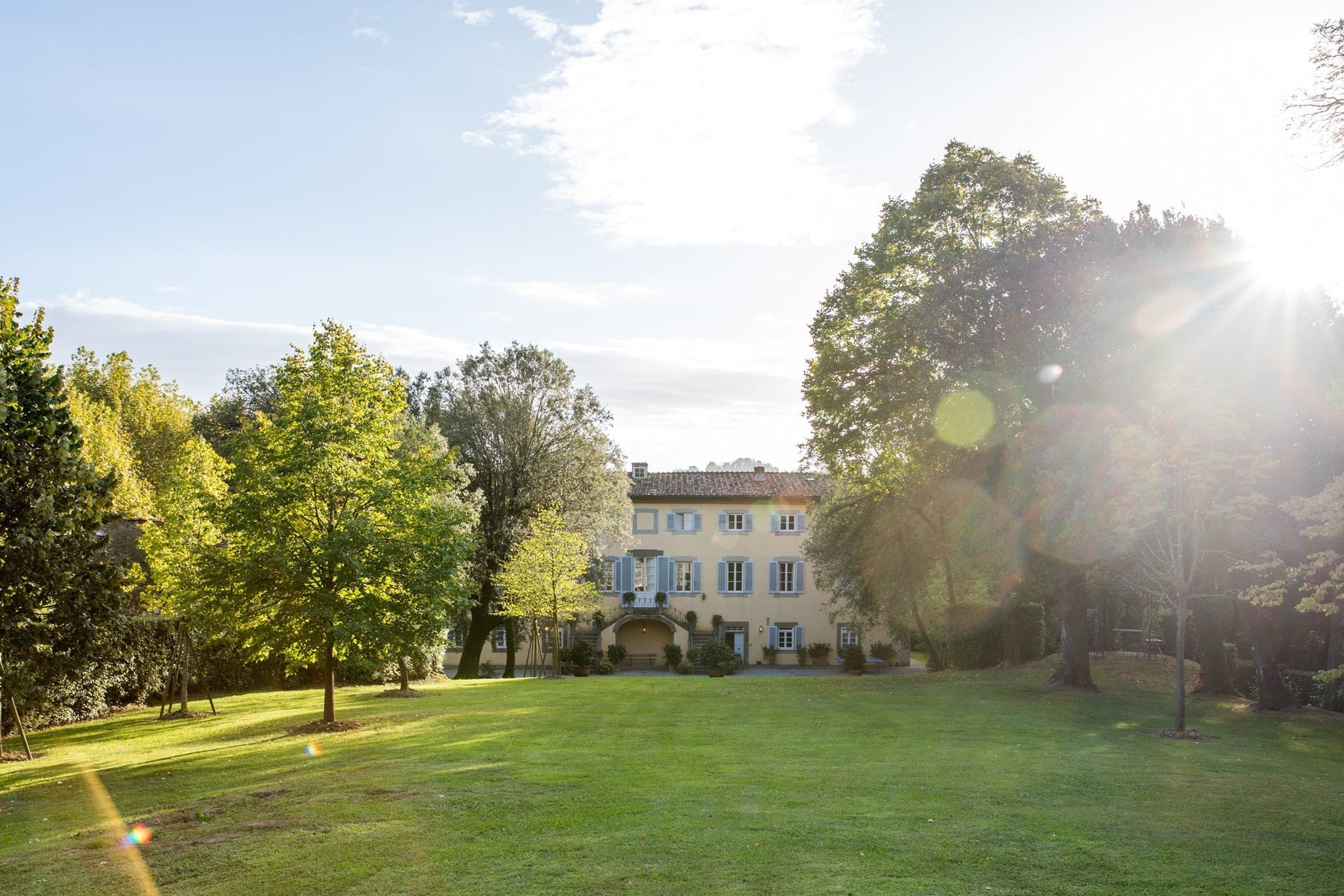 Three-story yellow villa with blue shutters beyond a grassy lawn and trees, sun flare at right