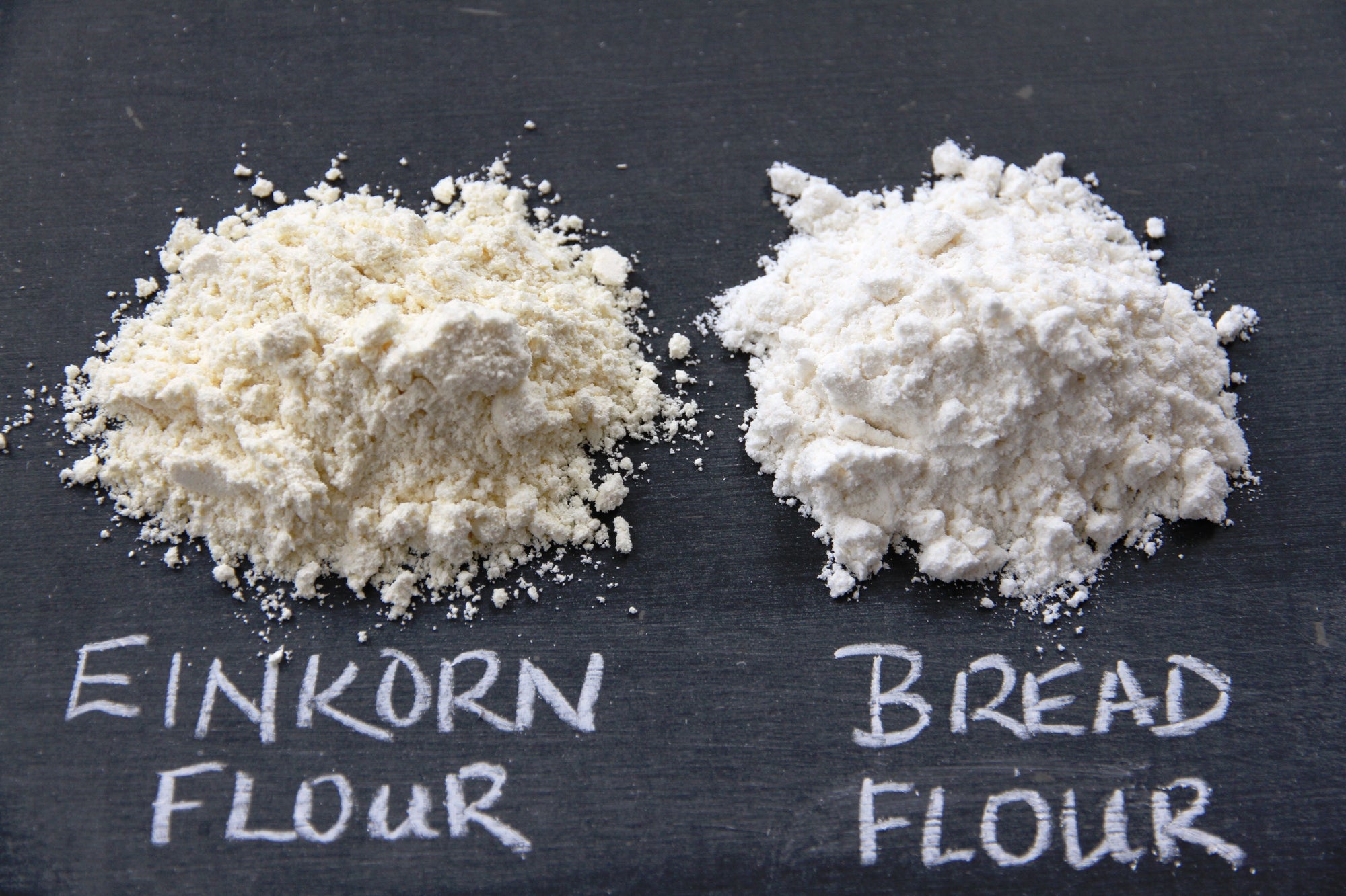 Two flour piles on chalkboard labeled 'EINKORN FLOUR' (left) and 'BREAD FLOUR' (right)