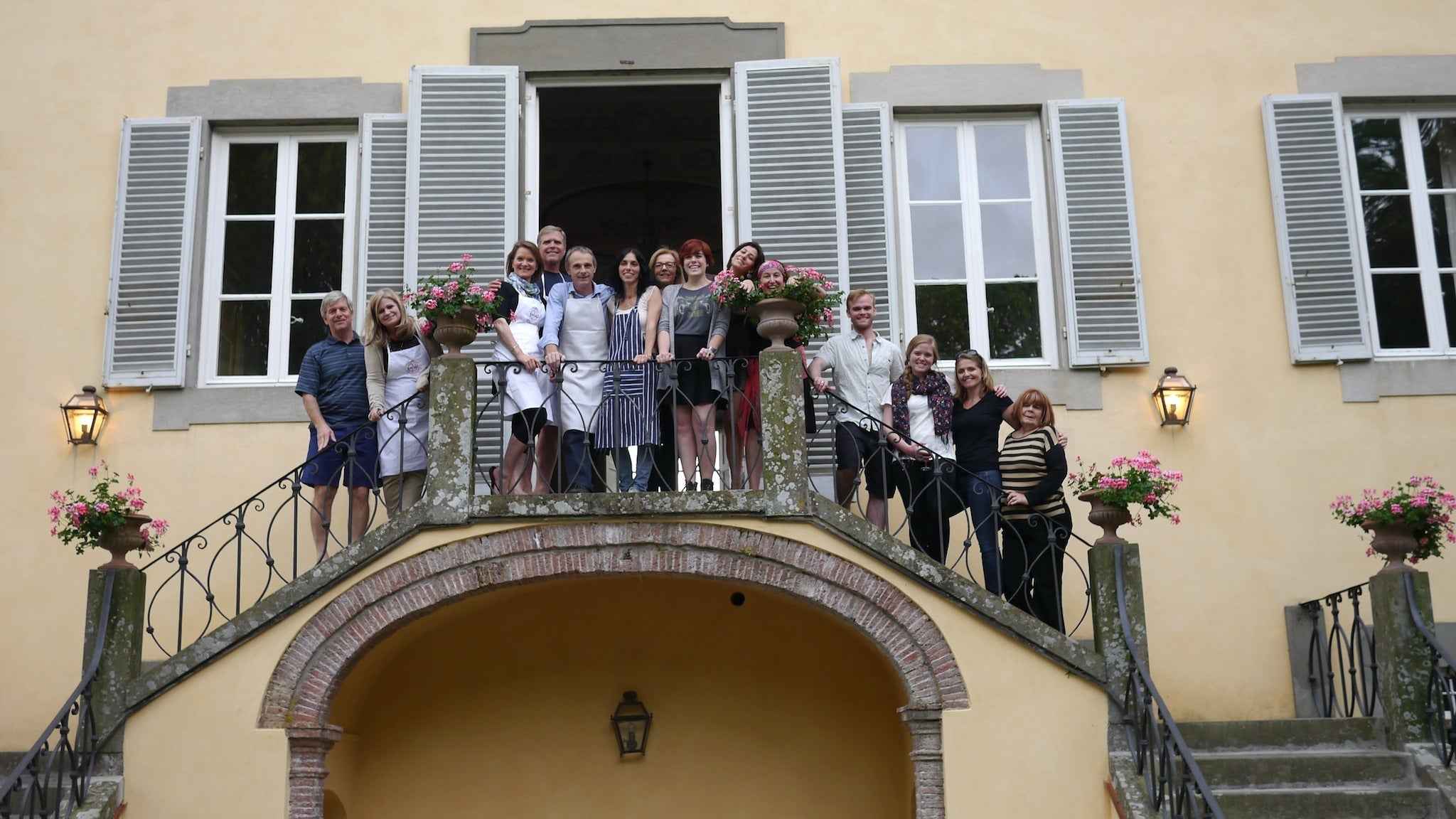 Group of people smiling together on a stone balcony outside a yellow house with open shutters and pink potted flowers