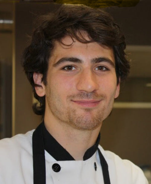 Young chef wearing a white coat with black collar, short dark hair and light stubble, smiling in a kitchen setting.