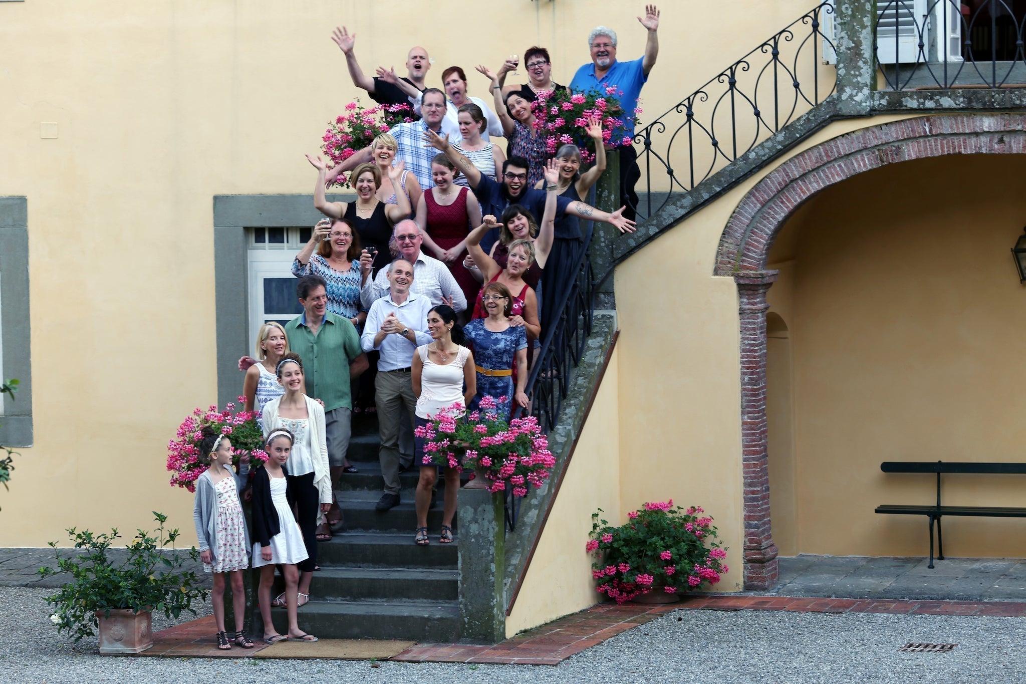 Large group of people posing on a stone staircase with pink geraniums, some waving, beside a yellow stucco building and arch.