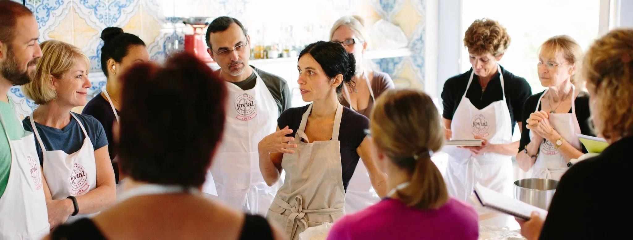 Cooking-class instructor leading students wearing Jovial aprons with visible text 'jovial' and 'LUCCA'.