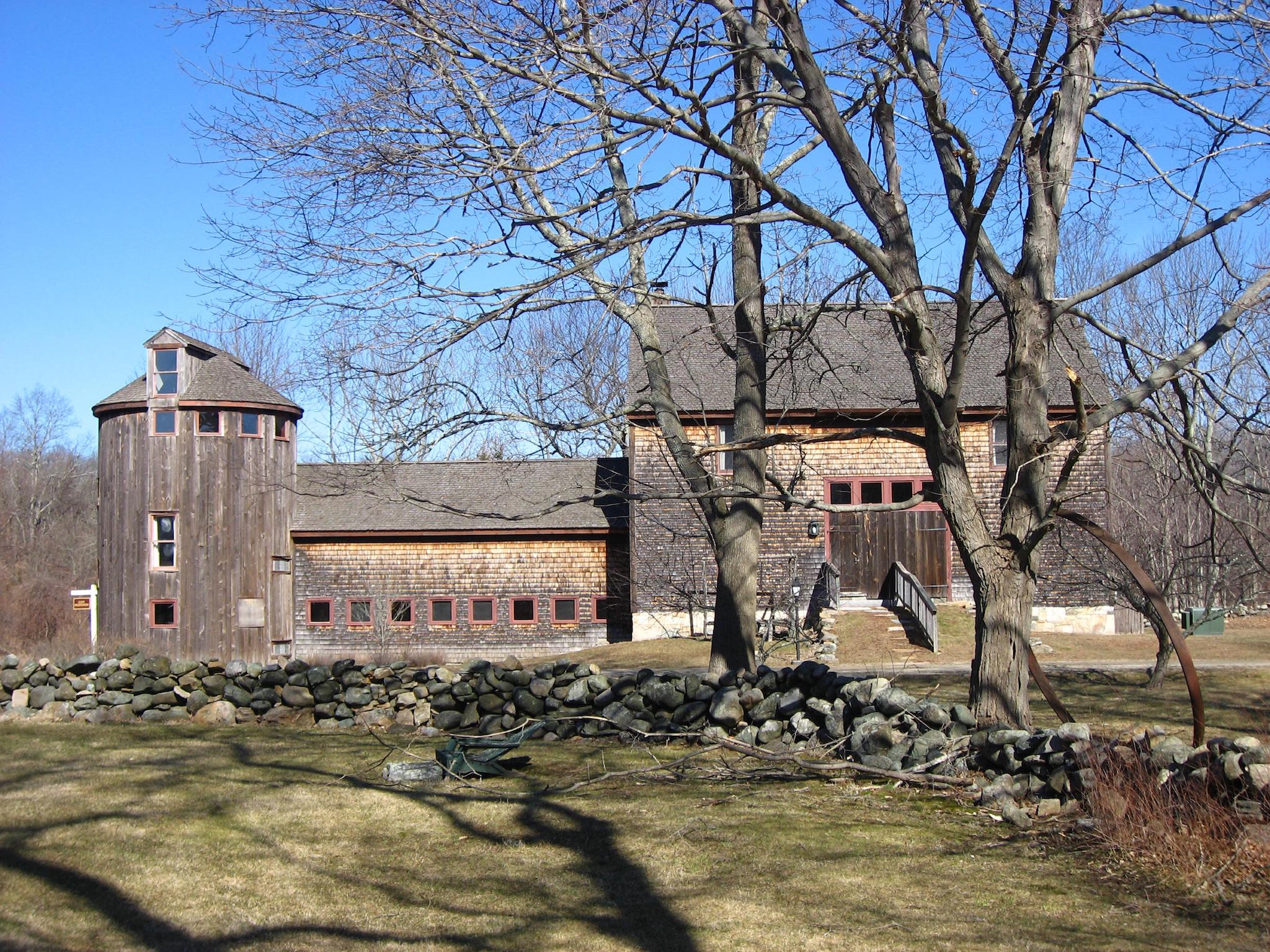 Wooden barn with round silo, low stone wall and leafless trees in foreground under a clear blue sky.