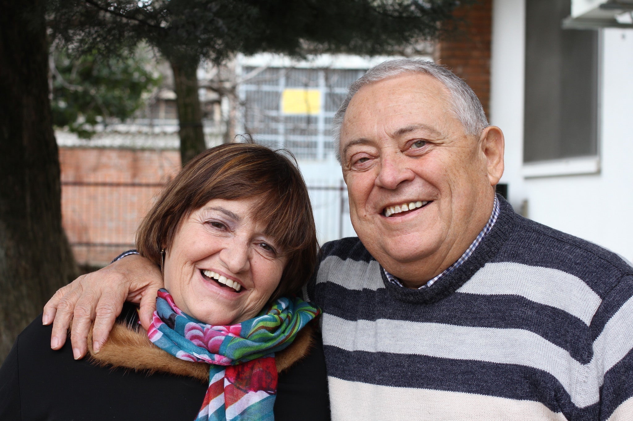 Smiling older couple outdoors, man with arm around the woman's shoulder; woman wears colorful scarf, man in striped sweater