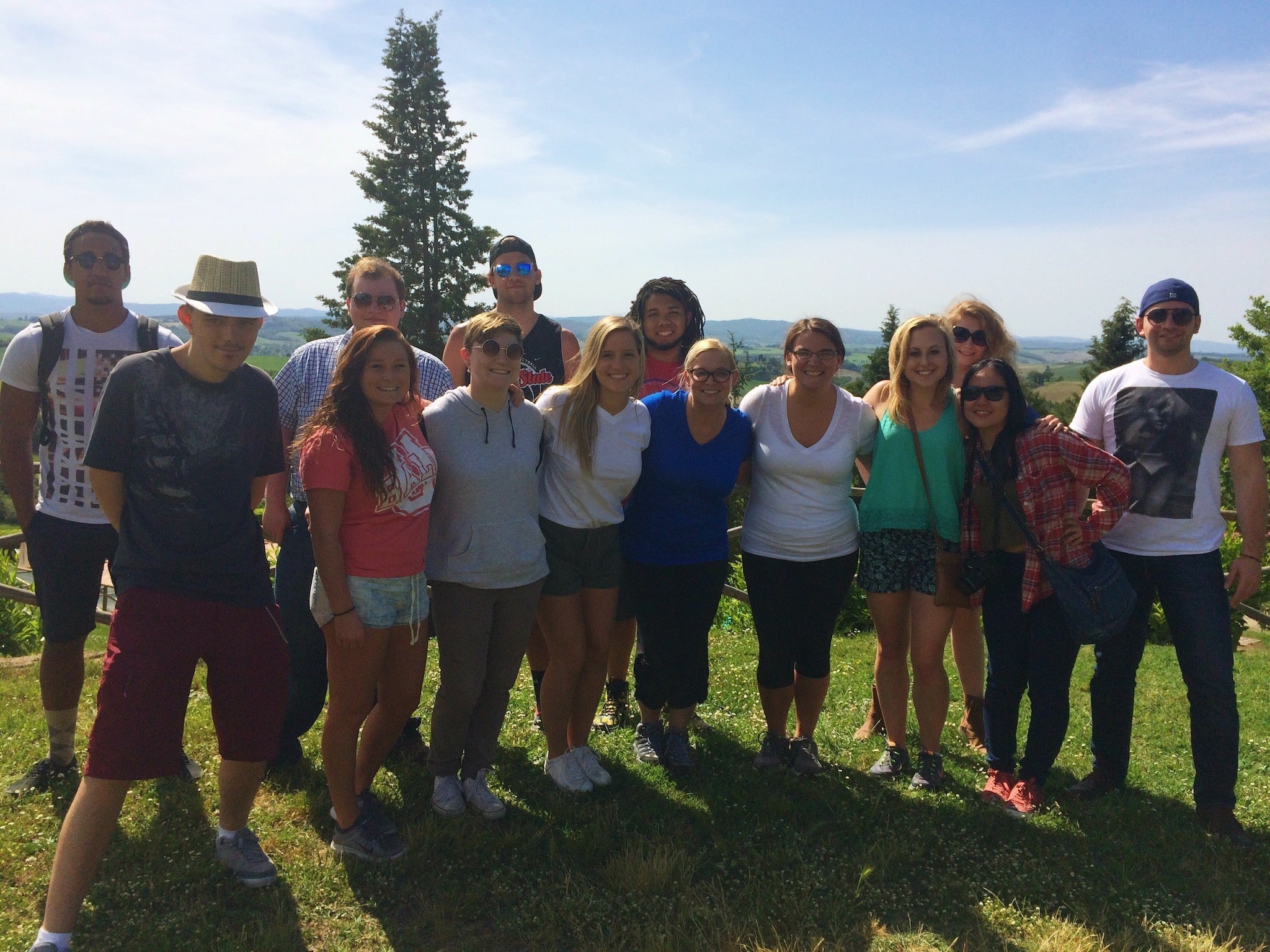 Thirteen people posing on a sunlit grassy hill with rolling countryside in the background, casual summer clothing.
