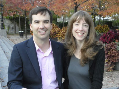 Smiling man and woman seated on stone steps with autumn trees and ornamental plants behind them.