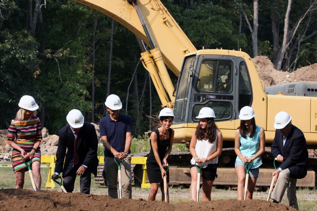 Seven people in hard hats shovel dirt at a groundbreaking ceremony in front of a yellow excavator