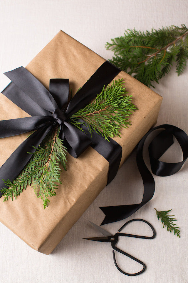 Brown kraft-wrapped gift with black satin ribbon and evergreen sprigs; black scissors and ribbon curl beside it.