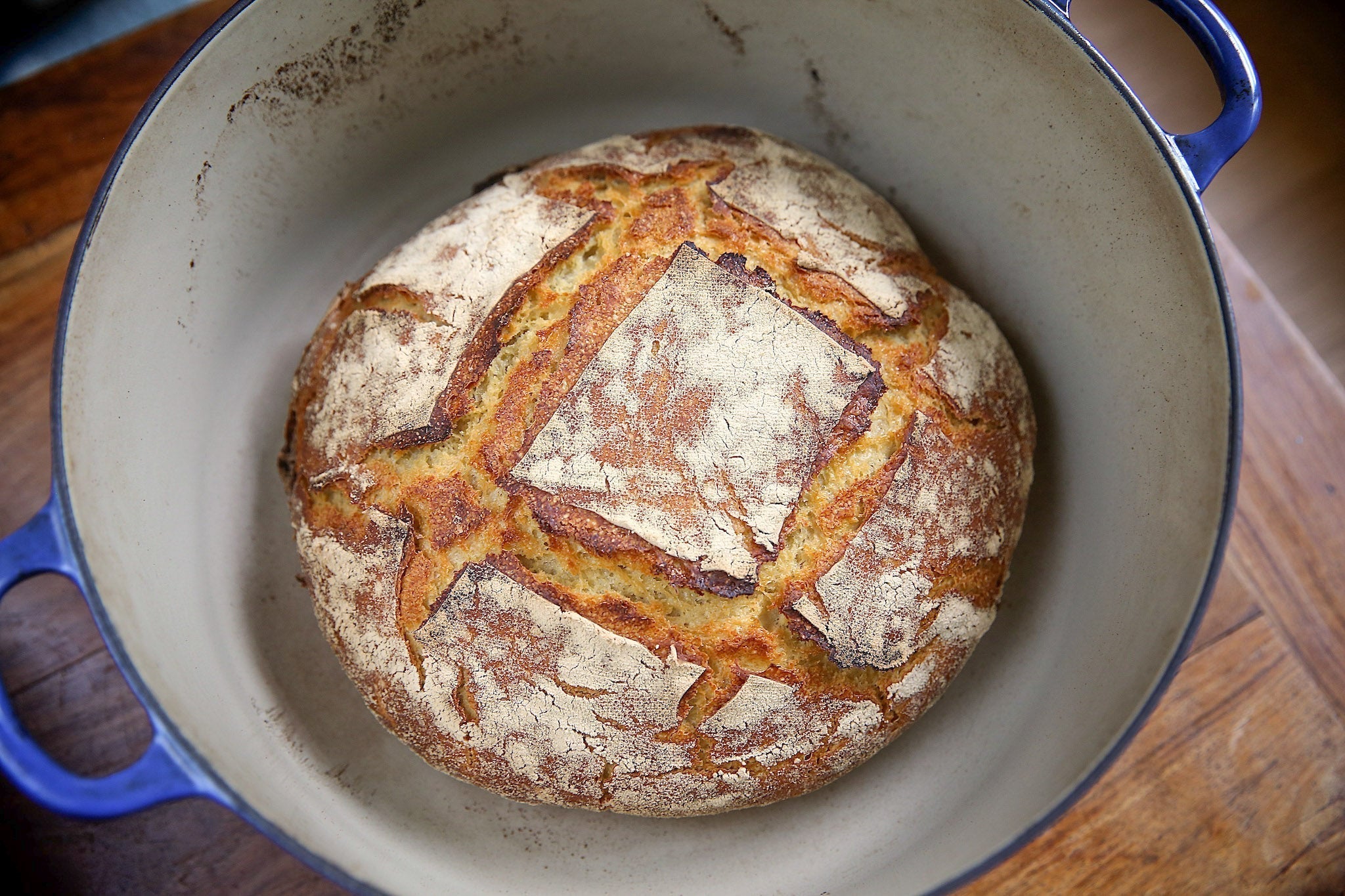 Round crusty artisanal sourdough loaf with scored square pattern, resting in a blue enameled Dutch oven.