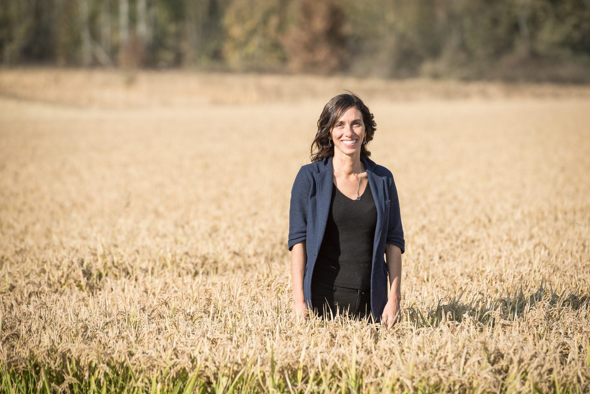 Woman standing waist-deep in a rice field, smiling, wearing navy blazer and black top.