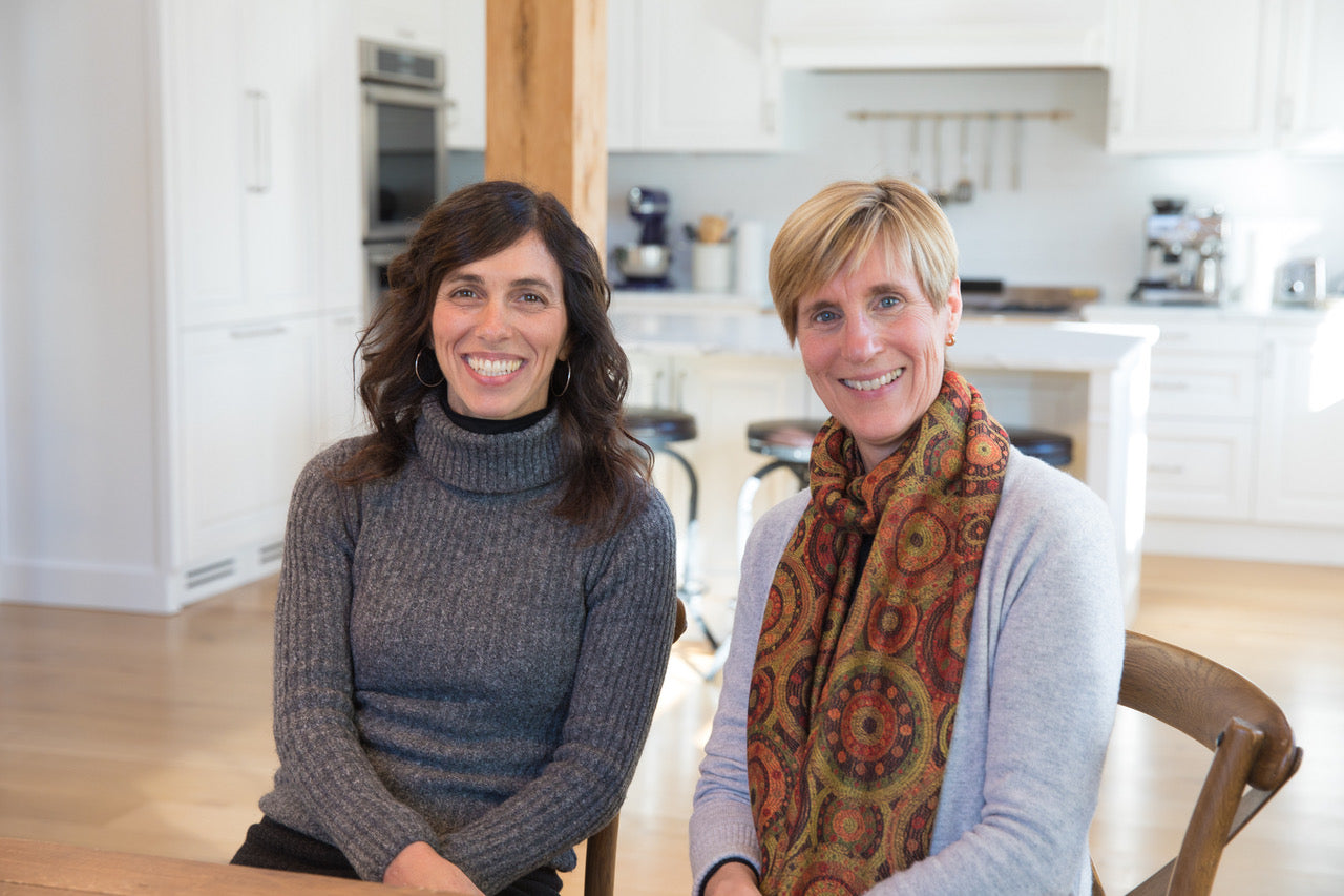 Two smiling women seated in a bright kitchen; left wears a gray turtleneck, right wears an orange patterned scarf.