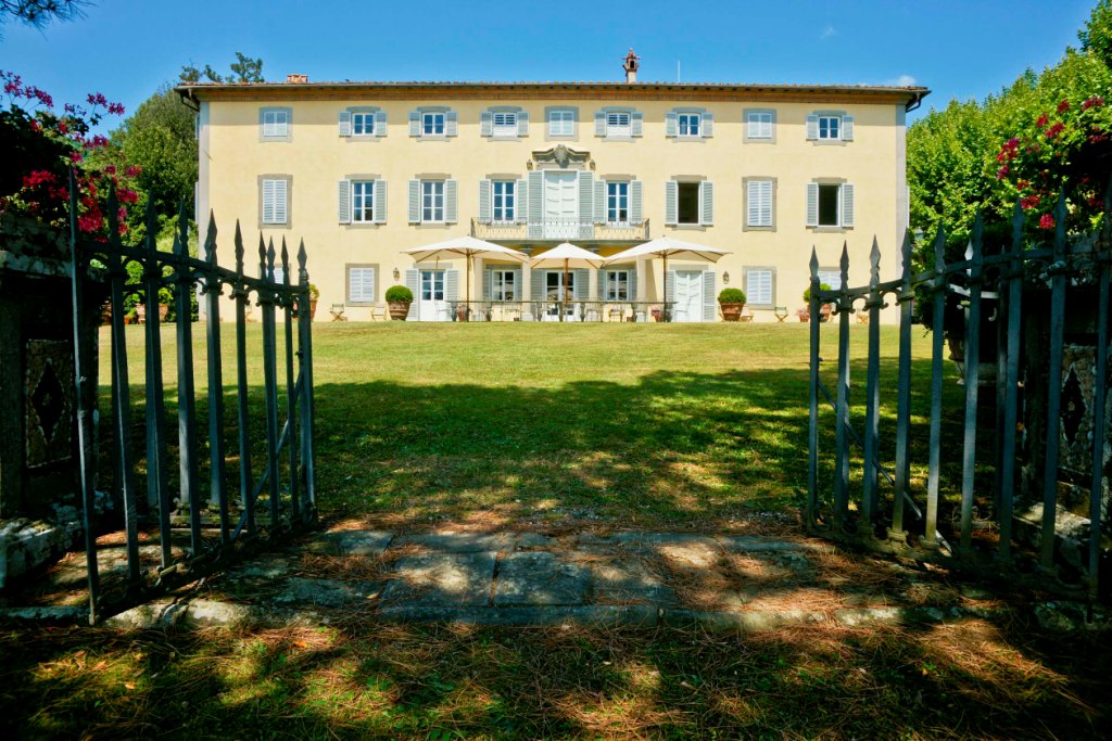 View through an open wrought-iron gate across a lawn to a sunlit Tuscan-style villa with white shutters and patio umbrellas.