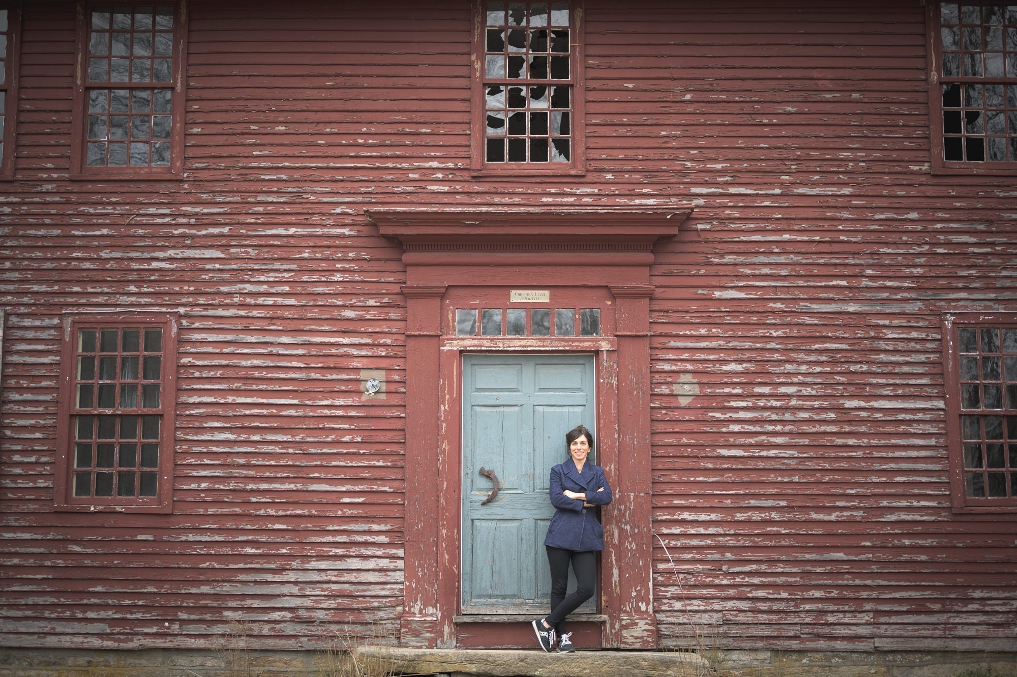 Woman leaning with arms crossed at a faded blue door set in a weathered red wooden building with peeling paint.