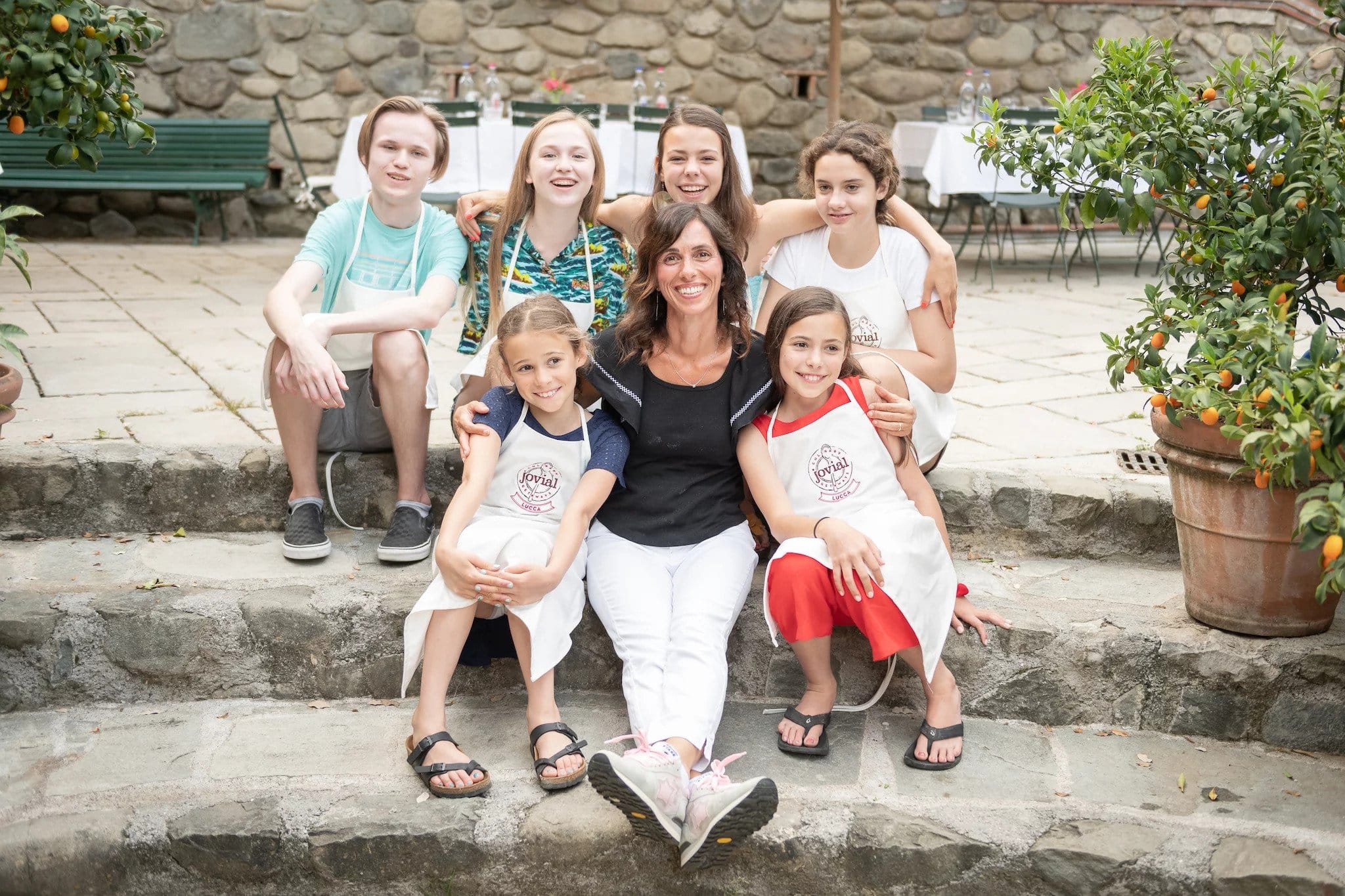 Smiling woman with children wearing Jovial Lucca aprons, seated on stone steps in a sunny courtyard with potted orange trees.