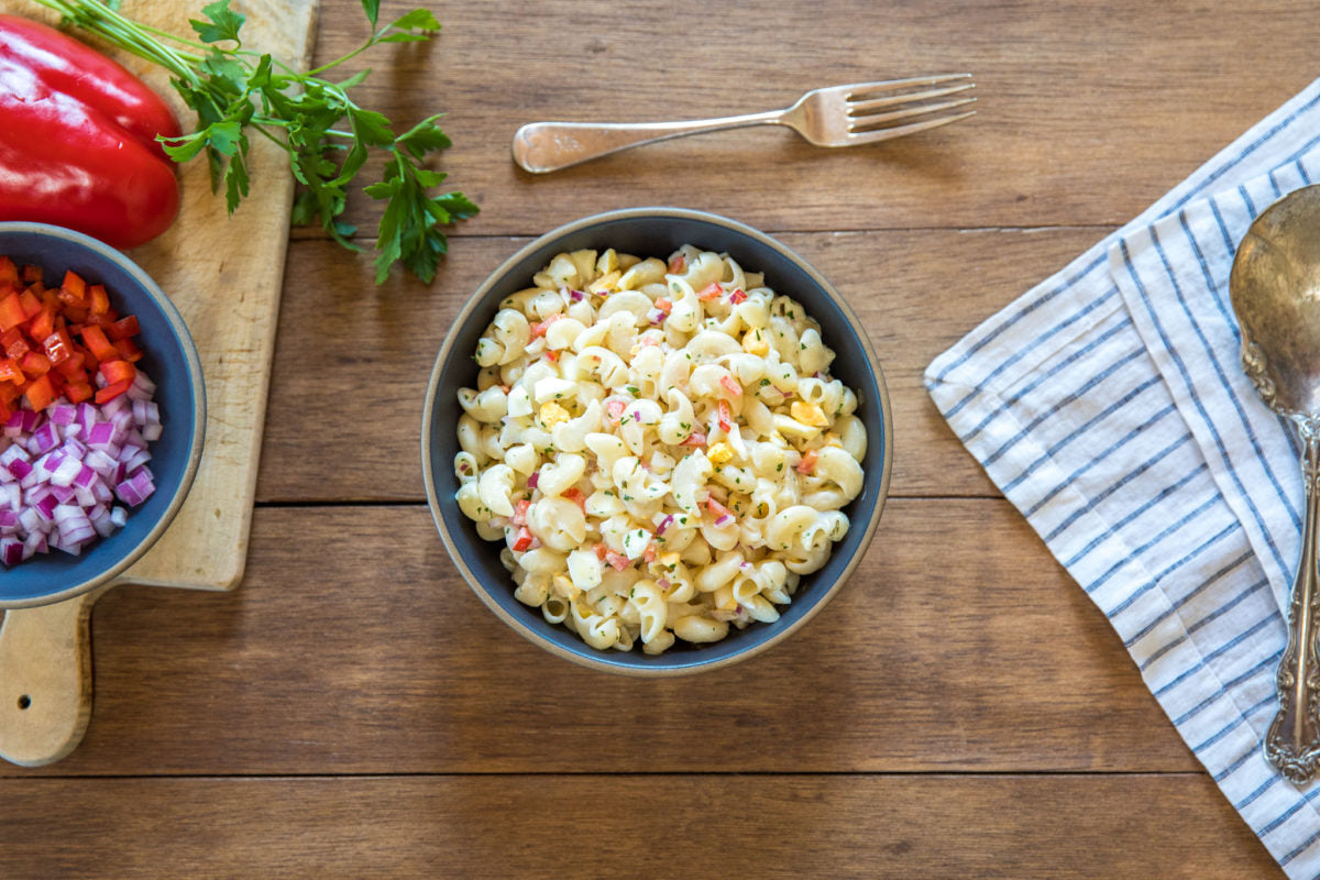 Bowl of gluten-free pasta salad with diced red pepper and red onion, fork and striped napkin on wooden table