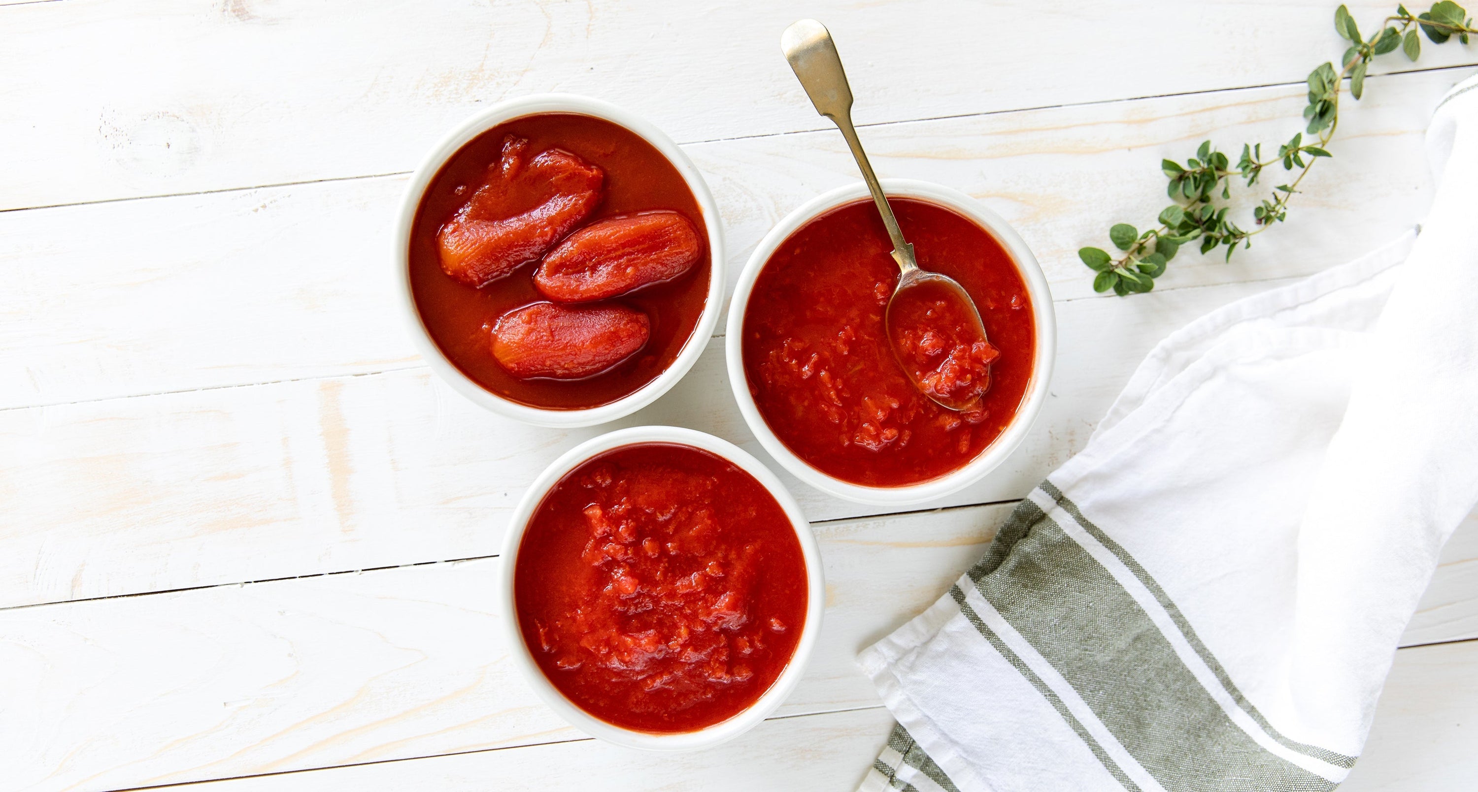Three bowls of canned tomatoes (whole peeled, crushed, diced) on a white wood board with a spoon and thyme