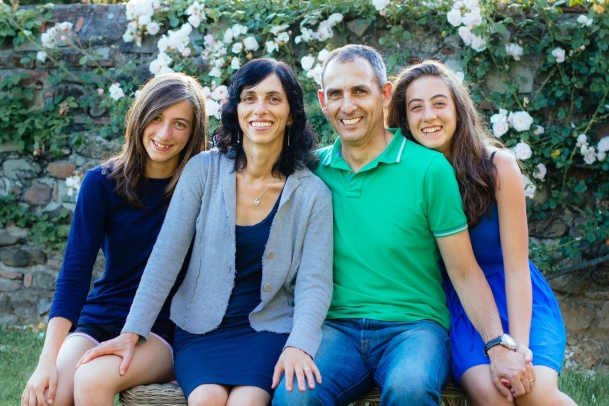Smiling family of four: Carla, Rodlofo and their two teenage daughters sitting on a bench in front of a stone wall covered with white roses.