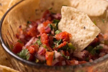 Bowl of salsa fresca with chopped tomatoes, red onion, cilantro and jalapeño, with a tortilla chip resting on top