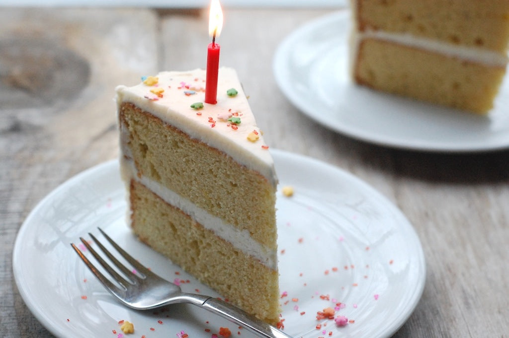 Three-quarter view of an Einkorn two-layer cake slice with white frosting, sprinkles, and a lit red candle on a white plate