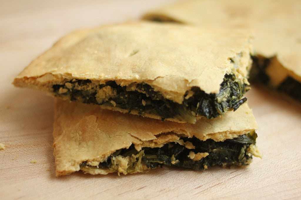 Close-up side view of mixed greens pie slice with flaky einkorn crust showing spinach and crumbly filling on a wooden board