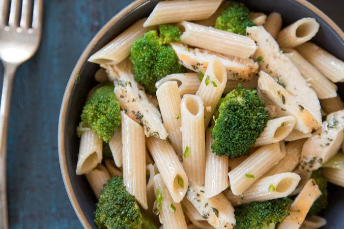 Bowl of brown rice penne with lemony broccoli and herbed chicken served in a shallow bowl on a dark blue surface