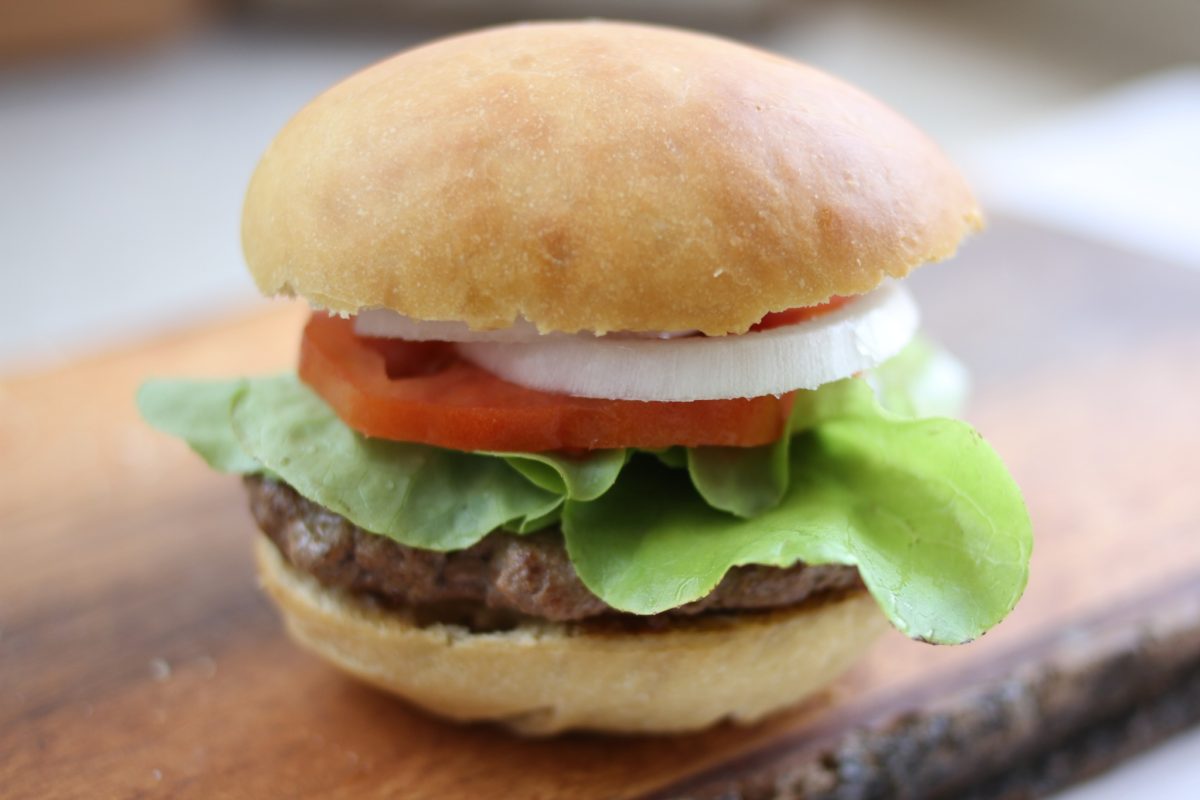Close up of sourdough einkorn hamburger bun topped with lettuce, tomato, and onion, showing golden crust on a wooden board