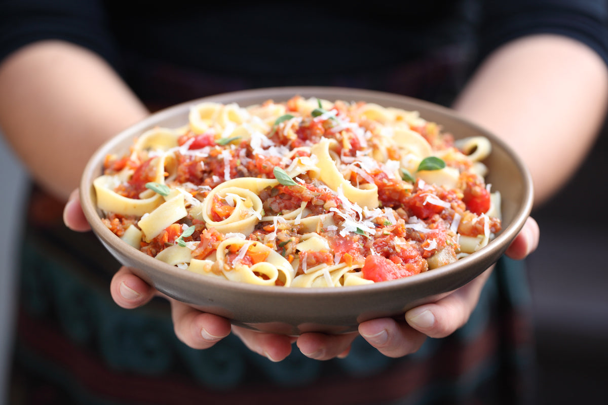 Close up of hands holding a bowl of brown rice tagliatelle topped with veggie bolognese, grated cheese, and fresh oregano
