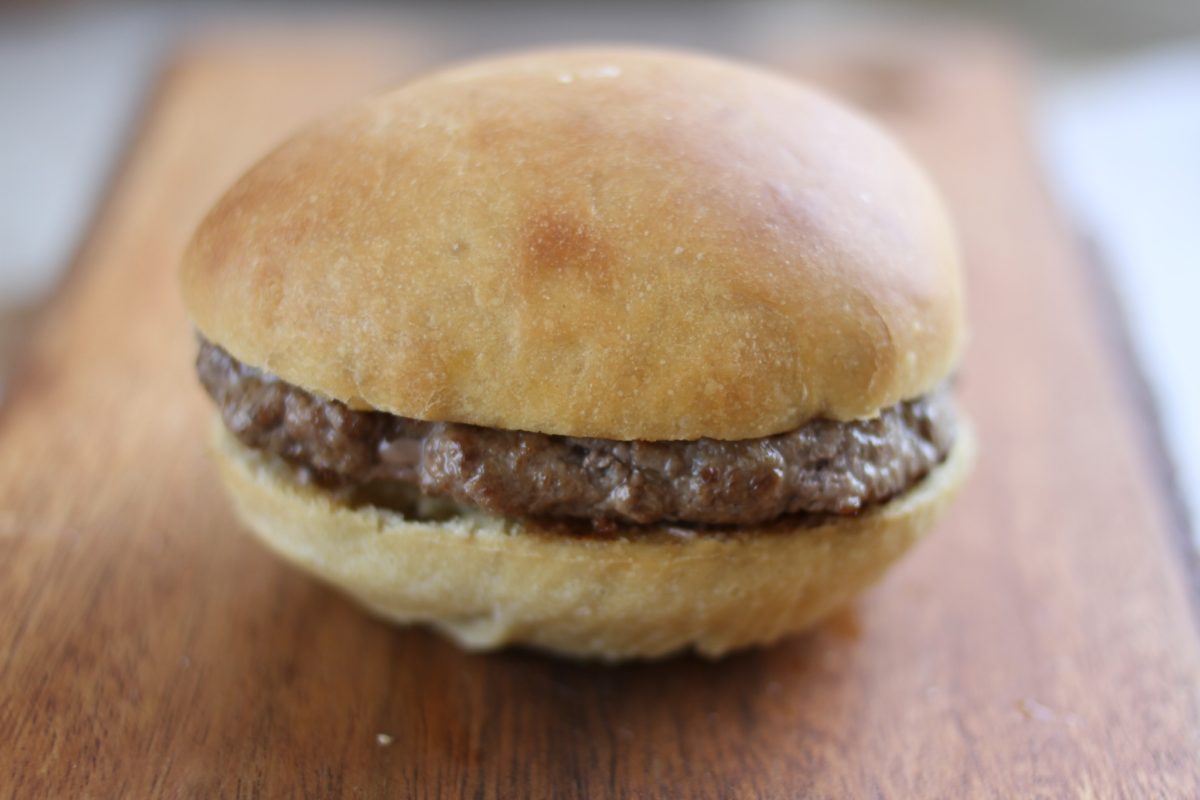 Close-up front view of an Einkorn hamburger roll showcasing a golden top and beef patty with a wooden cutting board background
