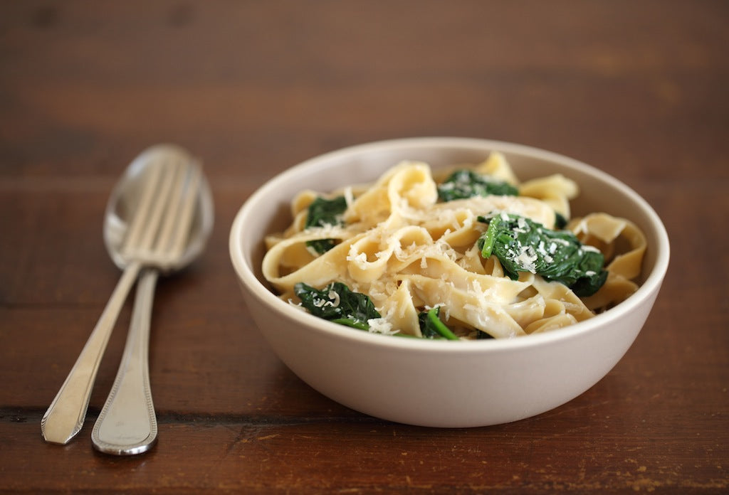 Bowl of brown rice tagliatelle with wilted spinach and cream sauce on a wooden table with a fork and spoon