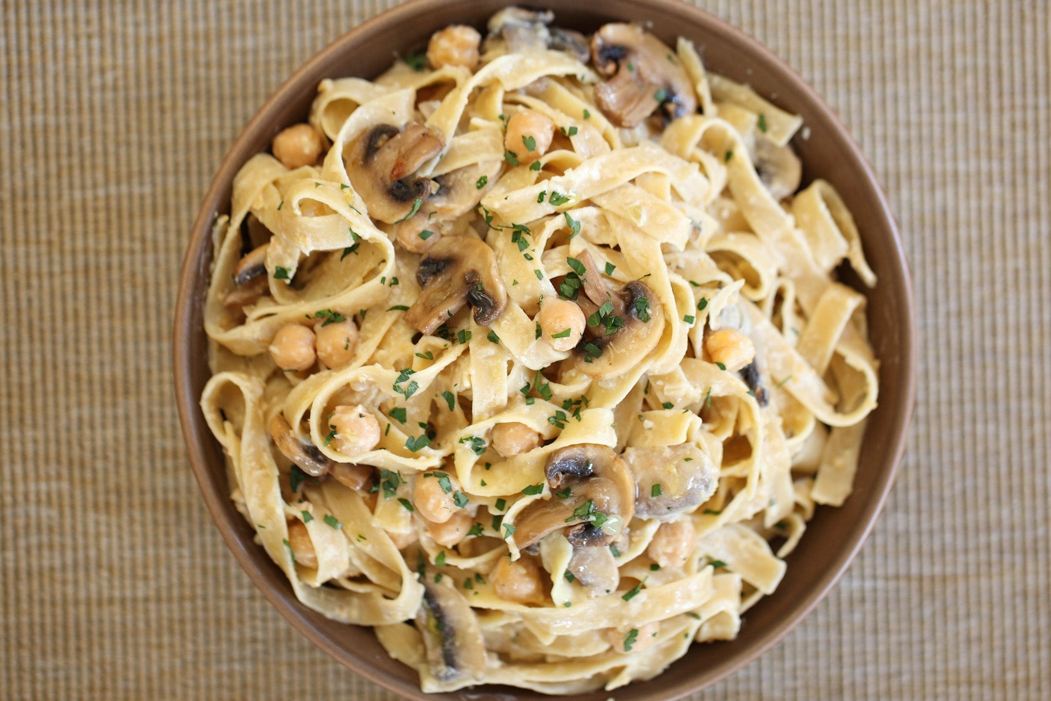 Top down view of a bowl of brown rice tagliatelle pasta tossed in creamy mushroom sauce with chickpeas on a woven placemat