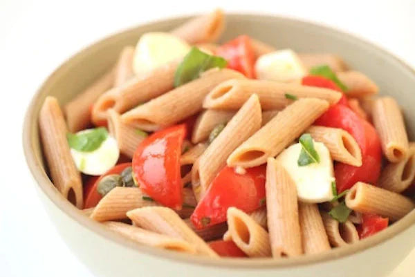 Bowl of penne pasta with cherry tomatoes, small mozzarella balls, basil and capers on a white background