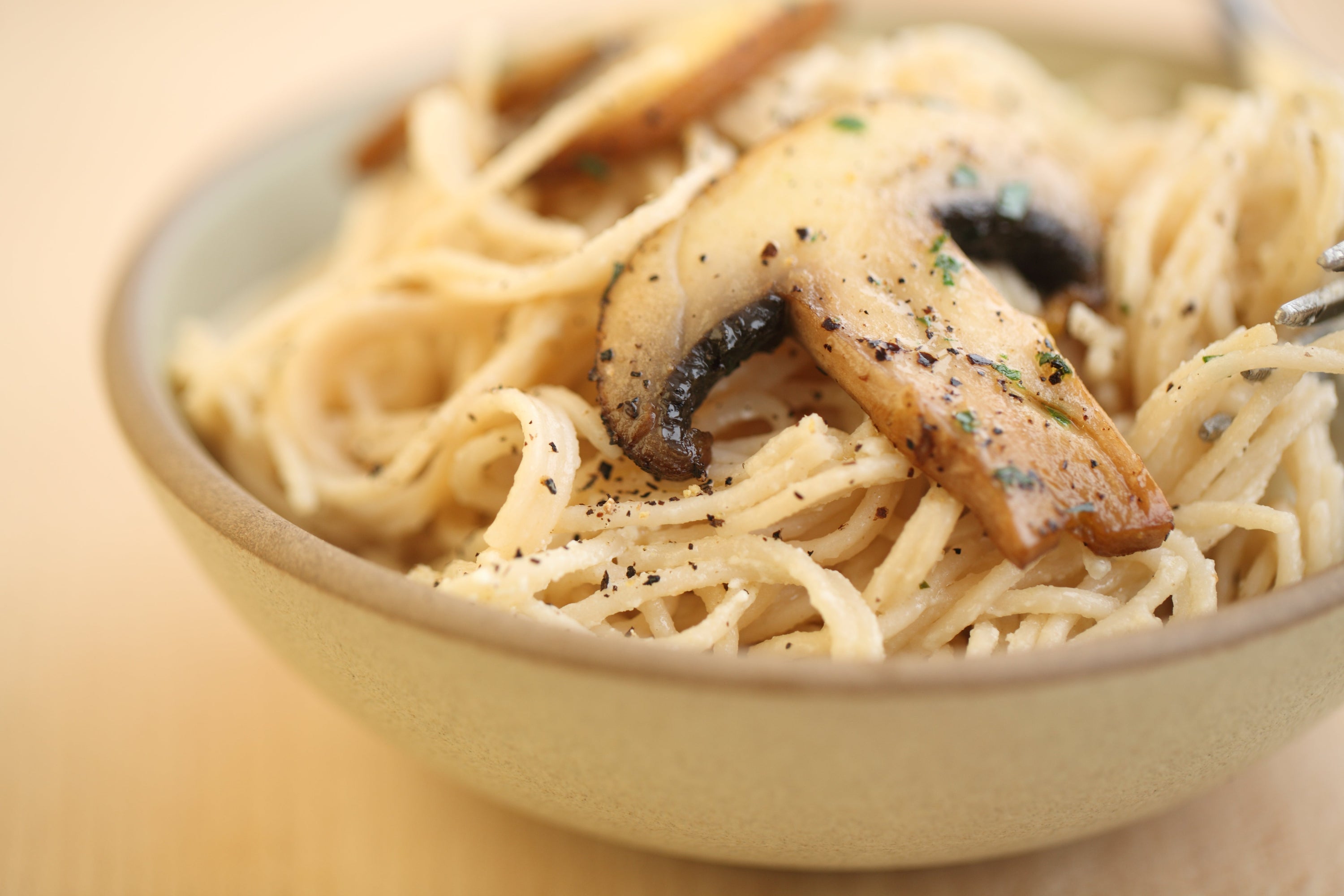 Close-up of capellini tossed with creamy goat cheese, sautéed mushrooms, and cracked black pepper in a ceramic bowl on a wooden table