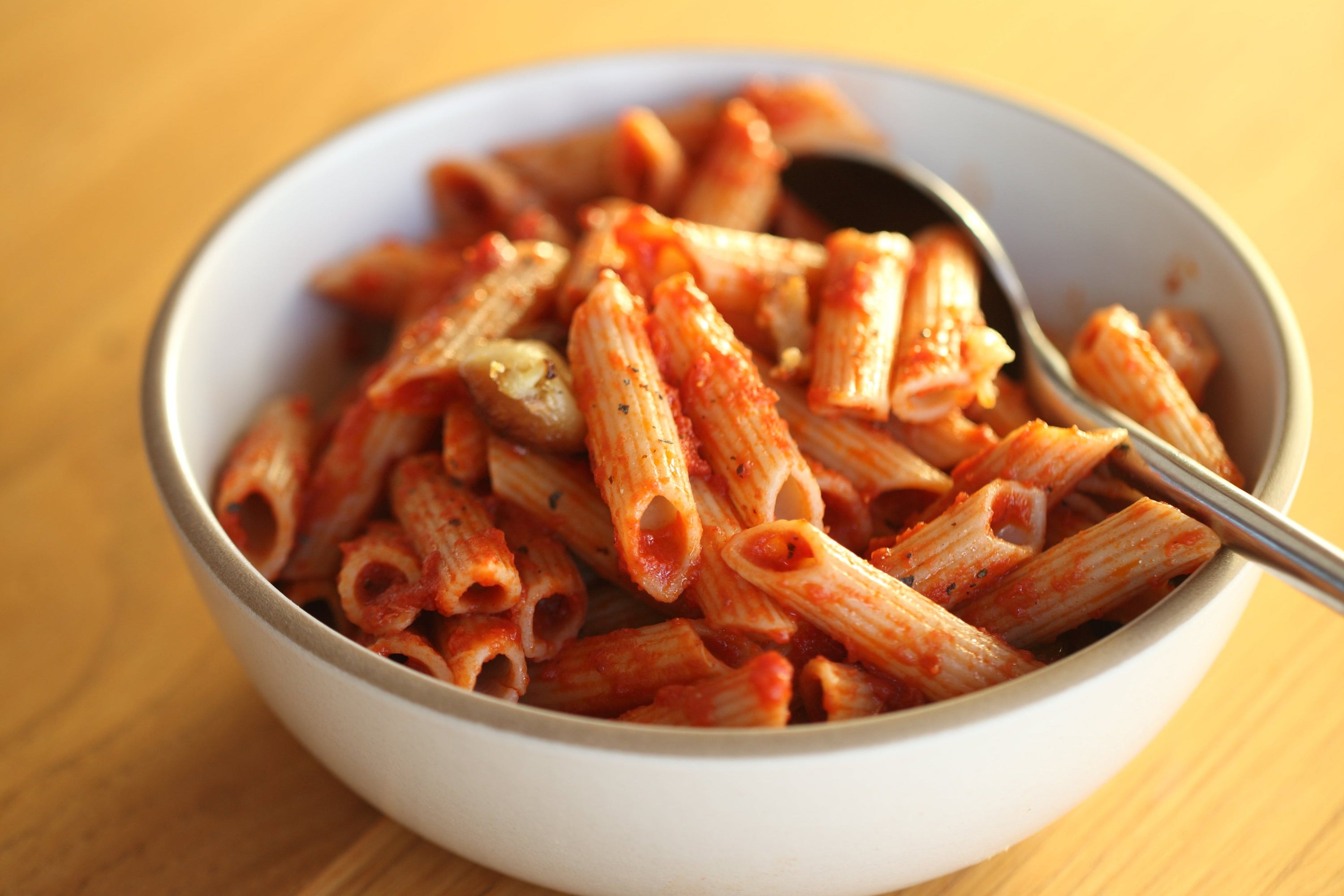 Close-up of penne tossed in roasted garlic and tomato sauce in a white bowl with a spoon on a wooden table