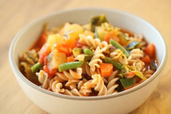 Close-up of bowl of minestrone soup showing fusilli pasta, green beans, carrots, and zucchini on a light wood surface
