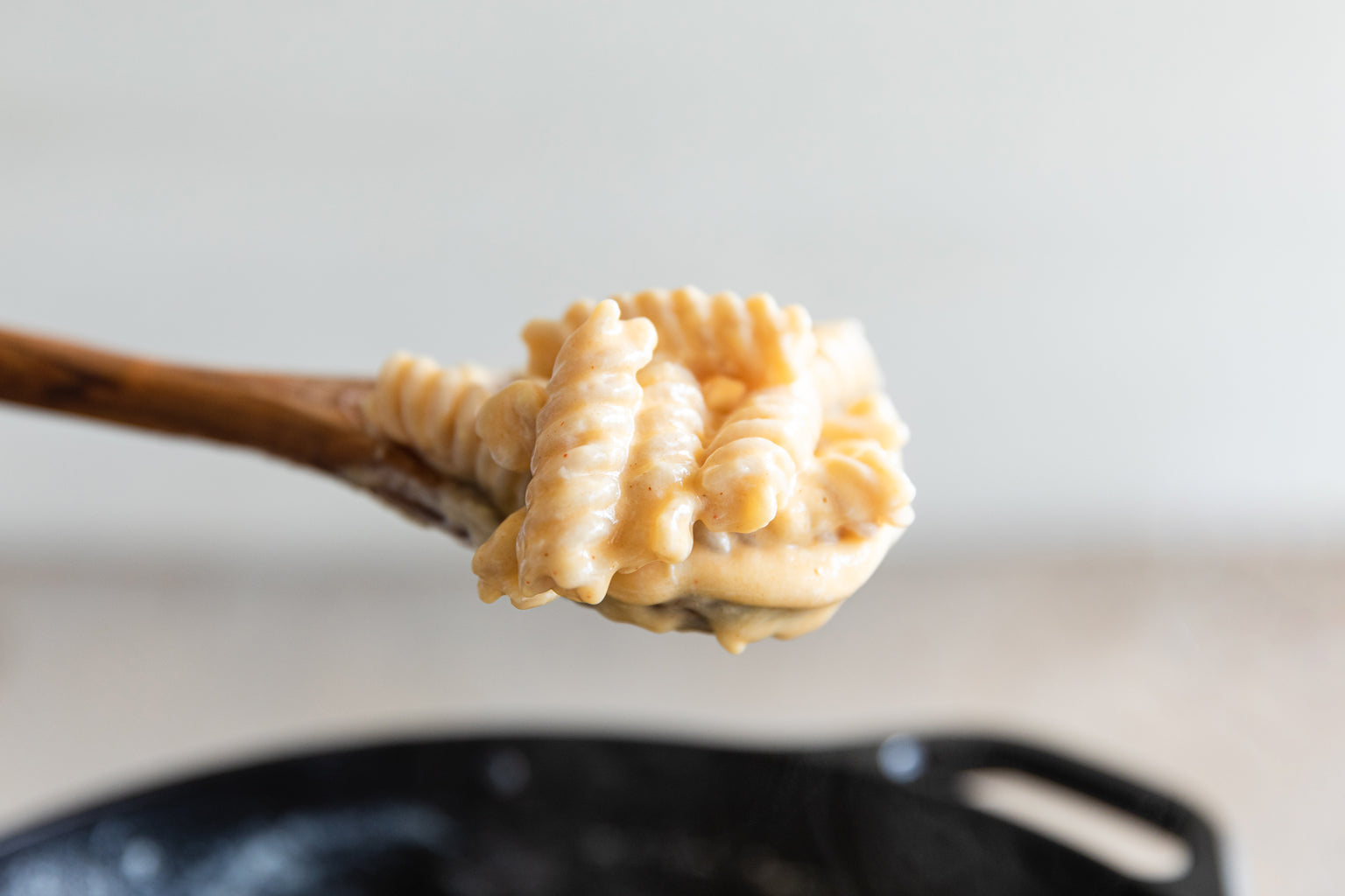 Wooden spoon lifting creamy mac and cheese made with gluten-free brown rice fusilli over a black skillet