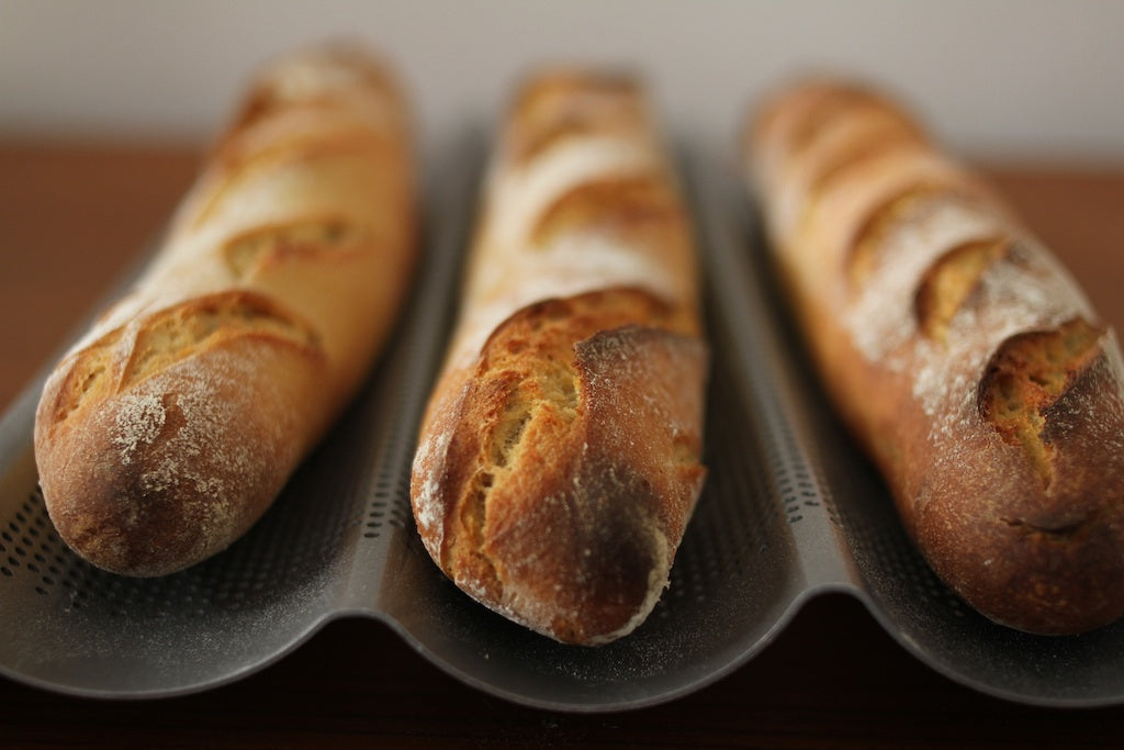Front close-up of three einkorn French baguettes showing golden crust and scored slashes on a ridged pan