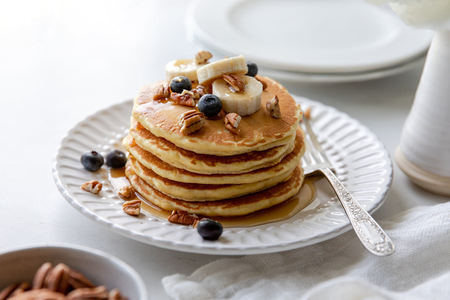 Stack of golden Einkorn pancakes topped with banana slices, blueberries, pecans, and maple syrup on a white plate