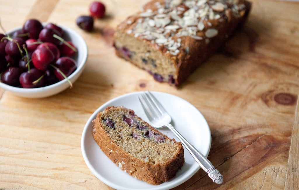 Slice of whole grain einkorn cherry almond quick bread on a white plate with a fork, loaf and cherries on a wooden board