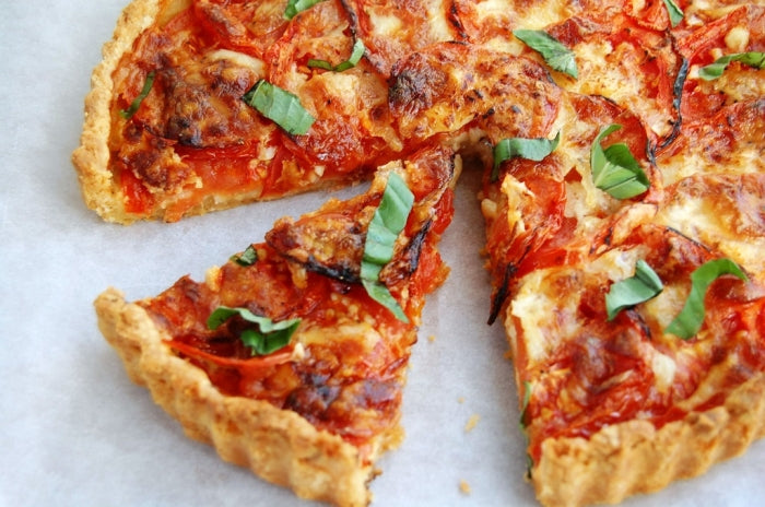 Close-up of tomato tart slice showcasing golden flaky crust, melted cheese, roasted tomato slices, and torn basil on parchment paper