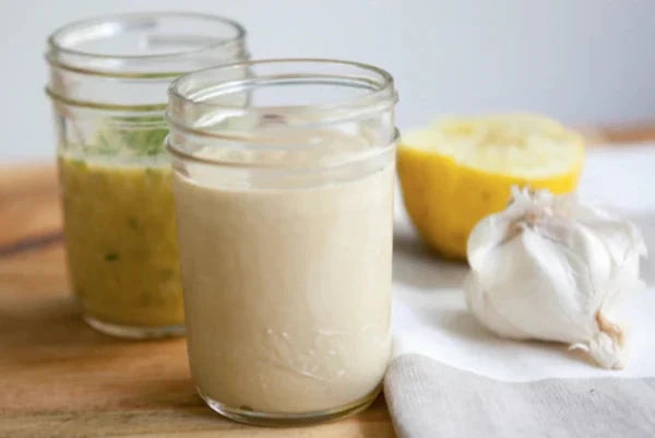 Two mason jars of lemon tahini dressing on a wooden board with a halved lemon and a head of garlic